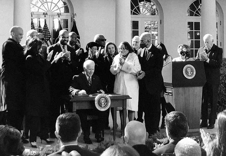 A black and white photo of President Biden seated at a desk in the Rose Garden surrounded by VP Harris and other leaders.