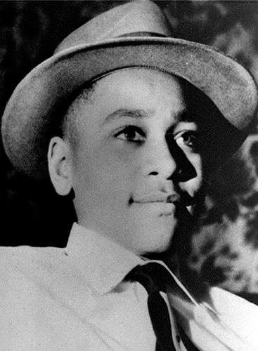 A black and white photo of Emmett Till. He's wearing a white button-up shirt, a black tie, and a grey hat.