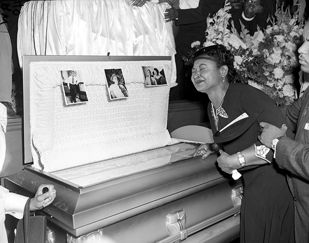 Mamie Till, mother of Emmett Till, weeps beside his open coffin. 3 pictures of Emmett are pinned to the lid of the coffin.
