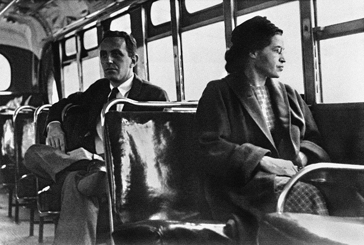 A black and white photo of Rosa Parks seated on a bus, looking out the window.