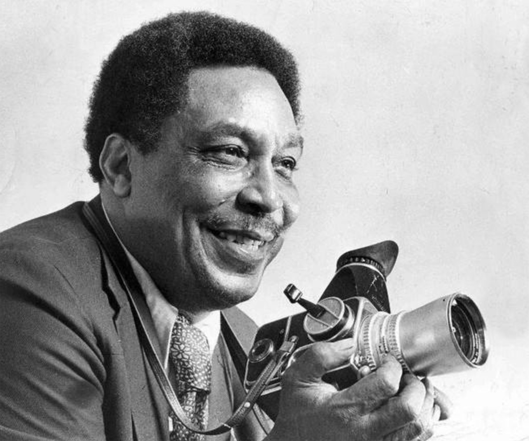 A black and white photo of Ernest Withers, a smiling man with a mustache and suit and tie. He’s holding a vintage camera. 